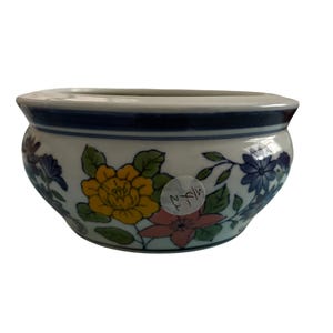 May include: A white ceramic planter with a blue rim and floral design. The planter features yellow, pink, and blue flowers with green leaves. The number "1714" is written on the side of the planter.