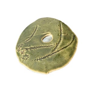 Könnte beinhalten: Handgefertigte, olivgrüne Keramikplatte mit einem zentralen Loch und strukturierten, blattartigen Mustern. Die Platte hat eine matte Oberfläche und ist mit sternförmigen Akzenten verziert.