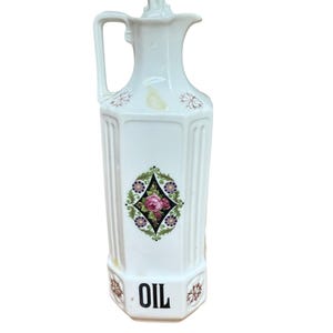 May include: White ceramic oil dispenser with a handle and a decorative floral design. The dispenser has a diamond-shaped floral pattern in the center and the word "OIL" in black letters at the bottom. The dispenser is approximately 10 inches tall.