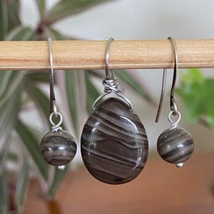 May include: A jewelry set featuring a teardrop pendant and matching earrings. The pendant and earrings are made of a brown and white striped stone, with silver-colored wire and hooks. The earrings are round and dangle.