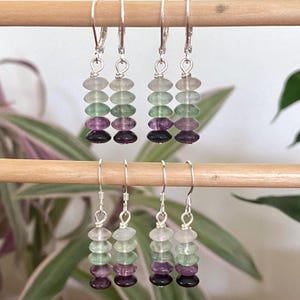 May include: Four pairs of dangle earrings featuring stacked, multi-colored gemstone beads in shades of clear, light green, dark green, and deep purple. Each earring is suspended from a silver-toned metal hook and wire finding, creating a vertical arrangement of polished, rounded stones.