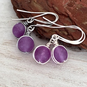 May include: A pair of handcrafted earrings featuring round, purple gemstones wrapped in silver wire. The earrings have a simple, elegant design with a hook closure. The gemstones are a vibrant shade of purple.