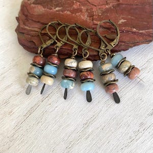 May include: A set of five stitch markers featuring antique bronze lever-back clasps. Each marker is adorned with a vertical arrangement of three small, smooth, multi-colored stone beads in shades of blue, beige, brown, and red, separated by bronze spacers.