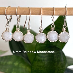 May include: Six pairs of silver wire-wrapped earrings with 8 mm rainbow moonstone beads. The earrings are hanging on a wooden dowel against a blurred green background. The text "8 mm Rainbow Moonstone" is visible below the earrings.
