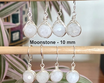 Puede incluir: Ocho pendientes de plata con piedras de luna redondas y facetadas de 10 mm. Cada piedra está asegurada en un engaste de alambre y cuelga de un gancho de plata. El texto Moonstone - 10 mm está impreso en el centro entre las dos filas.