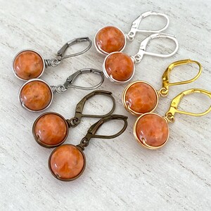 May include: Three pairs of dangle earrings featuring round, orange, mottled stone beads wrapped in wire. One pair has silver-tone leverback hooks, one pair has gold-tone leverback hooks, and one pair has antique bronze-tone leverback hooks.