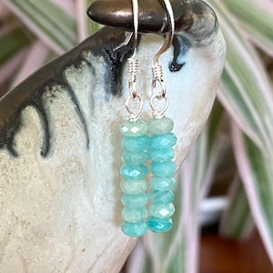 May include: A pair of dangle earrings featuring vertical columns of faceted, light turquoise-colored gemstone beads. Each earring is suspended from a silver-toned metal hook and includes a small silver-toned wire loop connecting the beads to the hook.