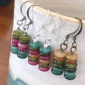 May include: Three pairs of dangle earrings featuring stacked, multi-colored gemstone rondelle beads. Each pair hangs from a different metal hook, including silver-toned and bronze-toned finishes. The beads display various shades of green, pink, and amber.