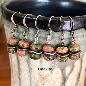 May include: Three pairs of dangle earrings featuring round, mottled pink and green Unakite beads separated by small metal spacers. The earrings hang from curved metal hooks in brass, silver, and copper tones, displaying the word Unakite below.