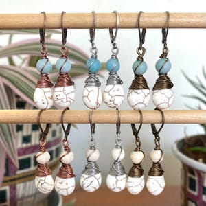 May include: A collection of handmade earrings displayed on a wooden dowel. Each earring features a white, cracked-stone teardrop bead, a light blue round bead, and wire wrapping in copper, silver, or bronze tones. The earrings have lever-back closures.
