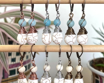 Puede incluir: Una colección de pendientes hechos a mano exhibidos en una varilla de madera. Cada pendiente presenta una cuenta de lágrima blanca agrietada, una cuenta redonda azul claro y un alambre enrollado en tonos cobre, plata o bronce. Los pendientes tienen cierres de palanca.
