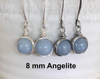 Puede incluir: Cuatro pares de pendientes de Angelita. Cada pendiente presenta una piedra redonda azul claro de 8 mm, envuelta en alambre de plata y suspendida de un gancho de plata. El texto "8 mm Angelita" se muestra debajo.