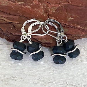 May include: A pair of dangle earrings featuring silver-toned leverback hooks and two faceted matte black beads separated by a curved silver-toned spacer on each side. The earrings hang from a silver-toned wire loop, showcasing a dark, textured design.