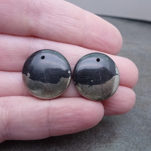 May include: Two round, dark gray pyrite gemstone beads with a metallic sheen and a small hole drilled at the top. The beads have a natural, textured appearance with a mix of dark gray and silver tones.