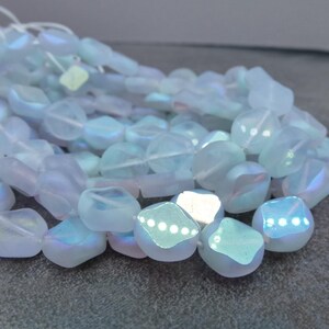 May include: A collection of iridescent, light blue beads with a frosted appearance. The beads are irregular in shape, with a mix of rounded and faceted surfaces. They are strung together on a white cord, suitable for jewelry making.