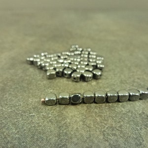 May include: A close-up of a pile of small, silver-colored, cube-shaped beads. A string of the same beads is strung on a wire.