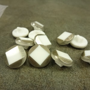 May include: A group of silver and white buttons with a square design. The buttons are arranged in a pile on a brown surface.