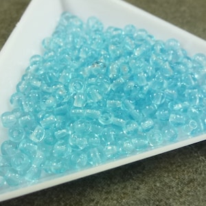 May include: A close-up shot of a white triangular dish filled with numerous small, translucent, light blue beads. The beads are cylindrical with rounded edges, creating a shimmering effect. The beads are likely for jewelry making or crafts.