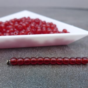 May include: A collection of small, translucent red beads. A string of the same red beads is strung on a gold-colored pin. A white triangular dish holds a larger pile of the beads, suggesting crafting supplies.