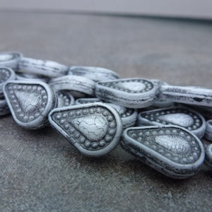 May include: A strand of white and gray teardrop-shaped beads with a textured surface and raised details. The beads are strung together on a clear thread.