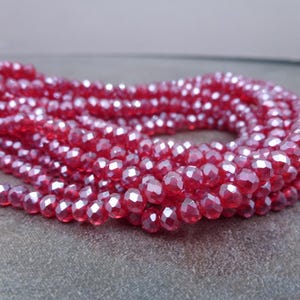 May include: A strand of small, round, faceted beads in a vibrant, translucent red color. Each bead features multiple flat surfaces that catch the light, creating a sparkling effect across the entire length of the coiled string of jewelry components.
