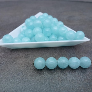 May include: A pile of light blue round beads in a white triangular dish. A strand of the same beads is shown below the dish.