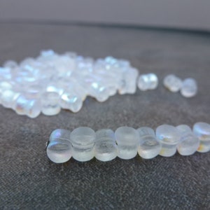 May include: A collection of iridescent, translucent glass beads. Some beads are strung on a thin wire, while others are scattered in a pile. The beads have a rounded, cylindrical shape and reflect a rainbow of colors.