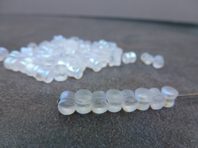 May include: A collection of iridescent, translucent glass beads. Some beads are strung on a thin wire, while others are scattered in a pile. The beads have a rounded, cylindrical shape and reflect a rainbow of colors.