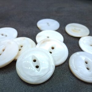 May include: A collection of ten white mother of pearl buttons with two holes each. The buttons are round and have a slightly raised edge.