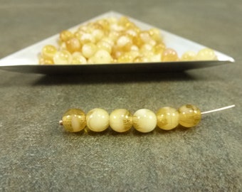 Puede incluir: Una hebra de cuentas de vidrio translúcidas de color amarillo y blanco con un brillo metálico. Las cuentas son redondas y tienen una ligera variación de tamaño.