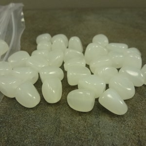 May include: A pile of white, opaque, smooth, oval-shaped beads. The beads are arranged in a loose pile on a gray surface.