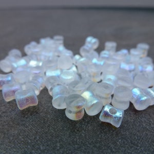 May include: A collection of small, translucent, square-shaped beads with a frosted appearance. The beads have a subtle iridescent sheen, reflecting colors like pink, purple, and blue. These beads are ideal for jewelry making and crafting.