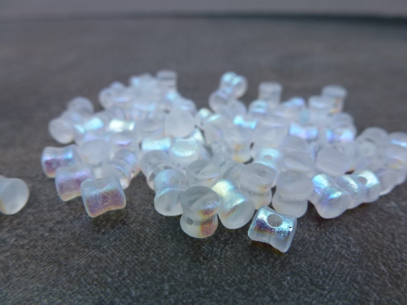 May include: A collection of small, translucent, square-shaped beads with a frosted appearance. The beads have a subtle iridescent sheen, reflecting colors like pink, purple, and blue. These beads are ideal for jewelry making and crafting.