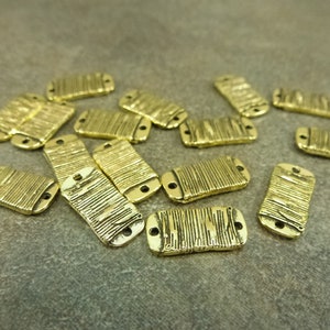 May include: A collection of small, rectangular gold-colored metal beads. Each bead has a textured, ridged surface and two small holes at each end. The beads are scattered on a neutral-toned surface.