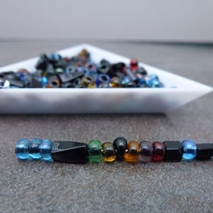 May include: A strand of colorful beads, including blue, green, black, and red, is displayed against a gray background. A white triangular dish filled with more beads sits in the background. The beads are of various shapes and sizes.
