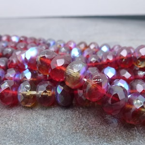 May include: Close-up of a strand of faceted beads. The beads are a deep red color with iridescent highlights of purple, blue, and gold. The beads are strung together, creating a shimmering effect. Ideal for jewelry making.