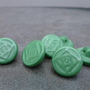 May include: A collection of six light green buttons. Each button is round with a raised geometric design in the center. The buttons are made of a smooth, glossy material and are scattered on a gray surface.