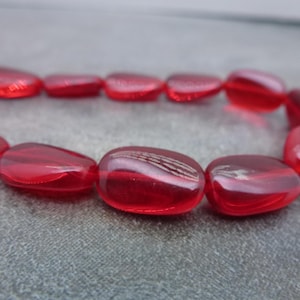 May include: A strand of translucent red beads. The beads are oval-shaped with rounded edges and a glossy finish. The beads are strung together, forming a curved shape against a gray background.