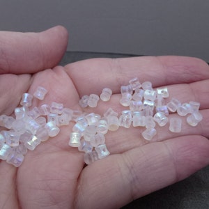 May include: A handful of iridescent, translucent glass beads. The beads are small, cylindrical, and have a subtle, shimmering effect. They are scattered across a neutral background, suggesting their use in jewelry making or crafts.