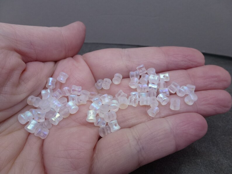 May include: A handful of iridescent, translucent glass beads. The beads are small, cylindrical, and have a subtle, shimmering effect. They are scattered across a neutral background, suggesting their use in jewelry making or crafts.