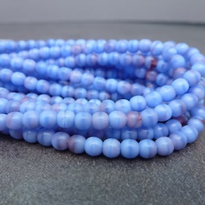 May include: Close-up of a bundle of light blue glass beads with a subtle pinkish-purple marbling effect. The beads are round and strung together, ideal for jewelry making or craft projects. The beads are approximately 6mm in diameter.