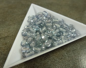 Puede incluir: Un montón de cuentas de vidrio azul claro en un plato triangular blanco. Las cuentas son de forma ovalada y tienen un acabado brillante.