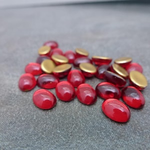 May include: A collection of red oval-shaped glass beads with a glossy finish, interspersed with gold-colored metal caps. The beads are scattered on a gray surface, creating a contrast between the colors and textures.