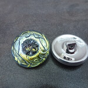 May include: A round, glass button with a silver metal back. The front of the button has a blue and green design with gold accents. The design features a stylized flower with a black center.