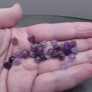 May include: A collection of small, round, purple amethyst cabochons. The cabochons are smooth and polished, with a slight sheen.