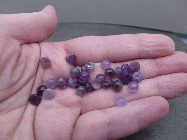 May include: A collection of small, round, purple amethyst cabochons. The cabochons are smooth and polished, with a slight sheen.