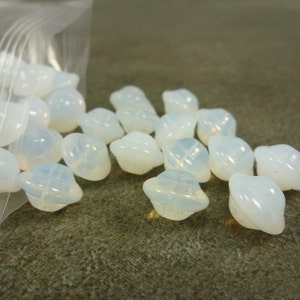 May include: A pile of white, opaque glass beads with a slightly iridescent finish. The beads are shaped like small, rounded, upside-down bowls.