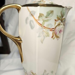 Puede incluir: Una jarra alta de porcelana blanca con asa y ribetes dorados. La jarra está decorada con rosas rosas y blancas pintadas a mano y hojas verdes. La tapa tiene un asa de anillo dorado. Una hermosa pieza para servir bebidas.