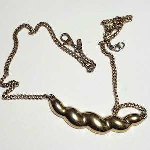 May include: A gold-toned necklace with a chain and a curved, textured pendant. The pendant has a series of connected, rounded shapes. The necklace is displayed on a white surface.