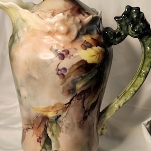以下が含まれることがあります： 柔らかいパステルカラーの手描きの花柄が特徴の、蓋付きのアンティーク磁器ピッチャー。ハンドルと蓋のディテールは緑色で、装飾的な葉のモチーフが付いています。ピッチャーの高さは約25cmです。